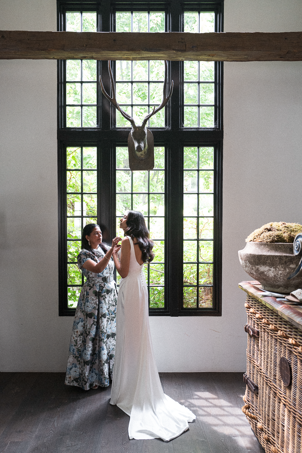 A woman in a white dress is being helped with her dress by another woman in a floral dress, inside a room with black window frames and greenery outside. A mounted deer head is on the wall above them, and a wicker piece of furniture with a large bowl 