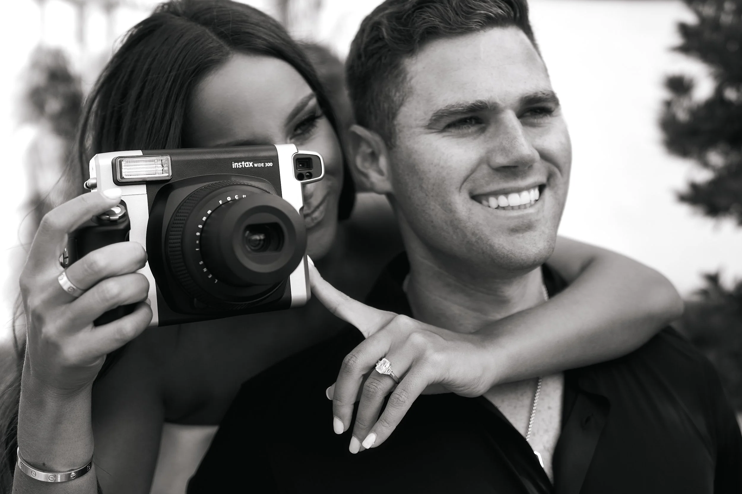 A woman taking a photo with a camera while a man stands beside her, smiling. The woman has her arm around the man's shoulders, and both are smiling, outdoors.