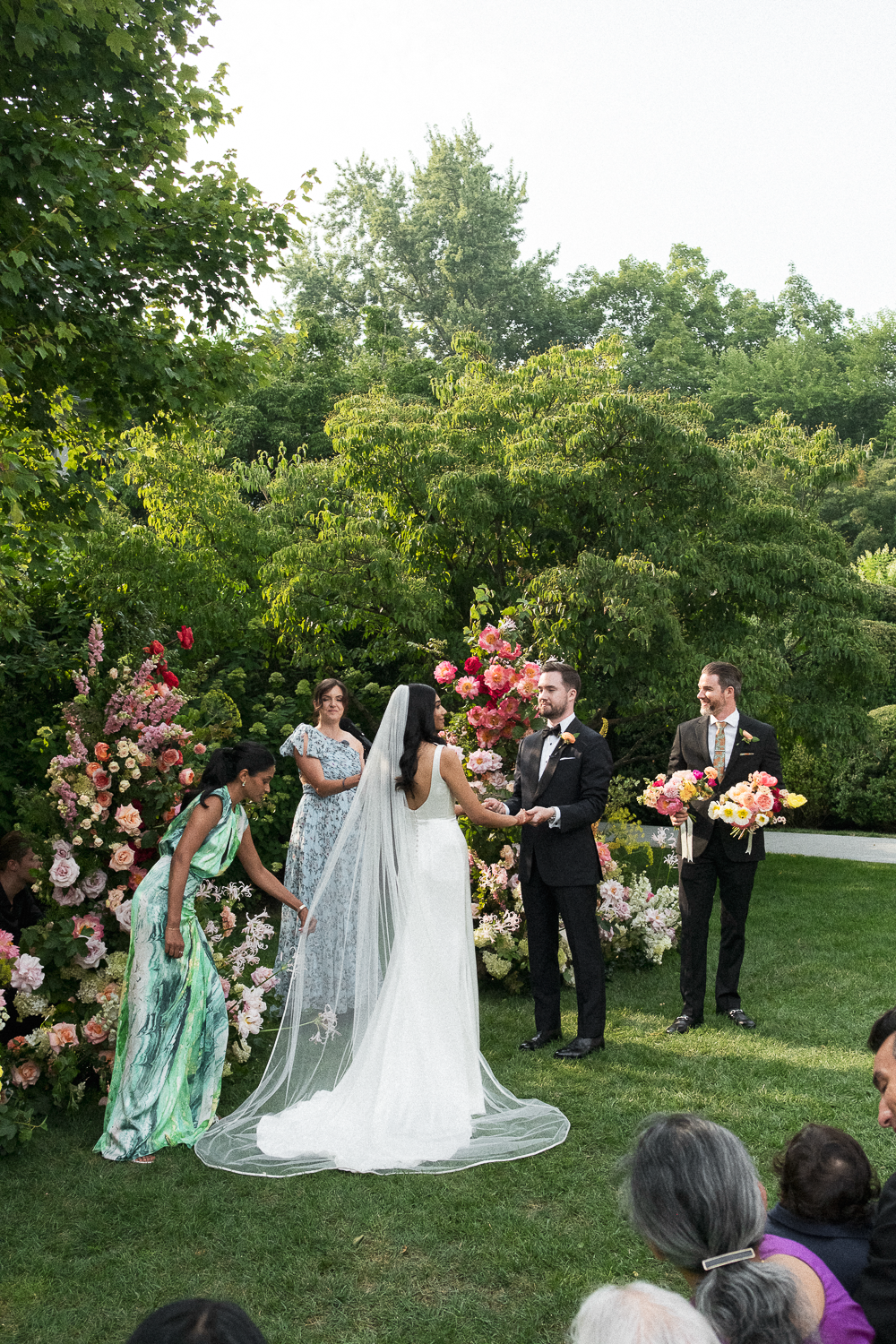 A wedding ceremony outdoors with a bride and groom exchanging vows, surrounded by colorful flowers and greenery, with guests watching.