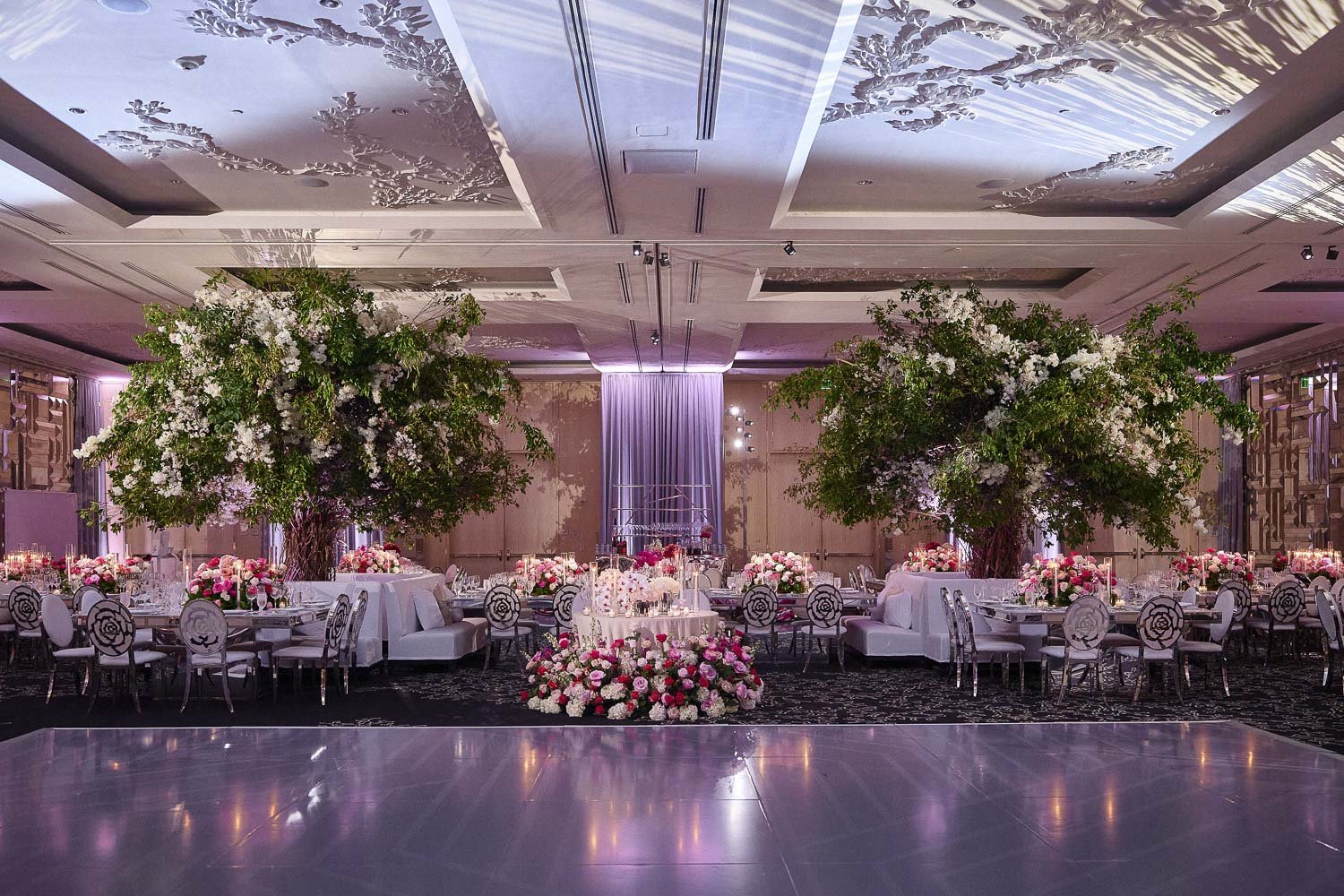 An elegant banquet hall decorated for a wedding reception with large floral trees, pink and white floral arrangements, and soft purple lighting.
