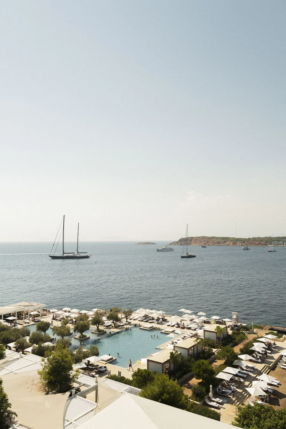 A seaside scene with a swimming pool and lounge chairs in the foreground, and sailboats on the water in the distance under a cloudy sky.