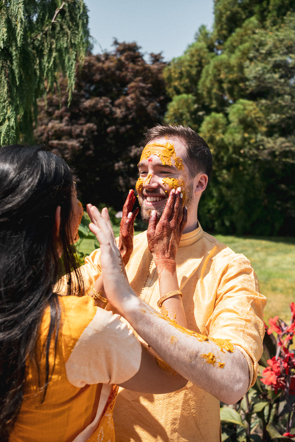 A man and woman celebrating together outdoors during a traditional Indian event, with the woman applying a yellow paste on the man's face, both smiling. The man is wearing a yellow traditional outfit, and the woman has henna on her hands. Trees and g