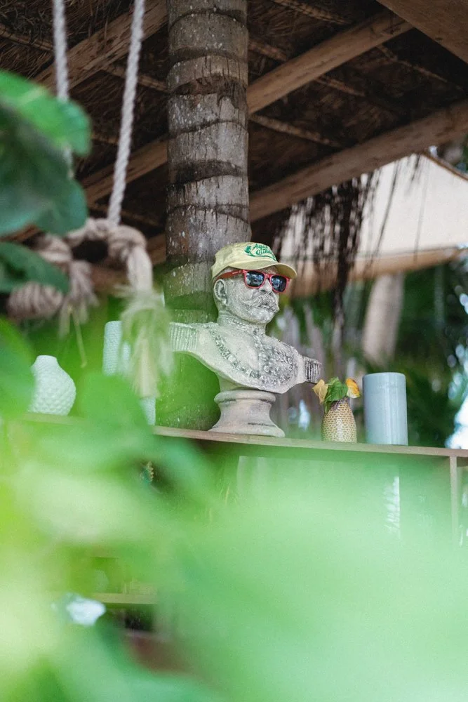 Bust of a man wearing sunglasses and a yellow cap, surrounded by decorative items on a shelf in a tropical setting with a thatched roof.