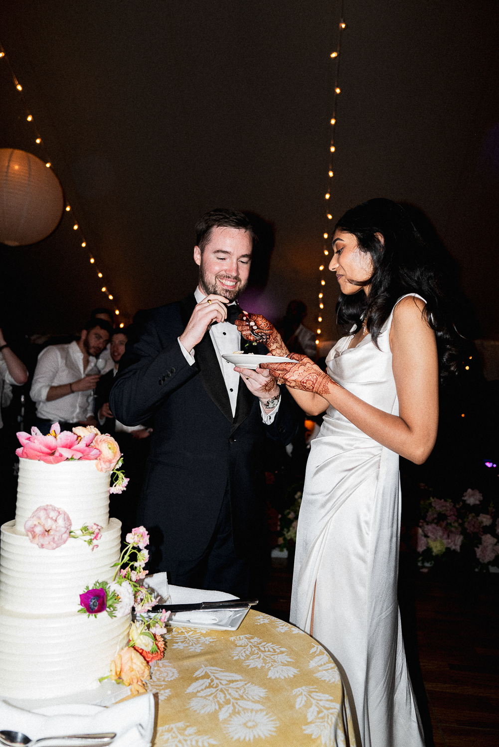 Bride and groom sharing wedding cake, with the bride using henna decorated hands.