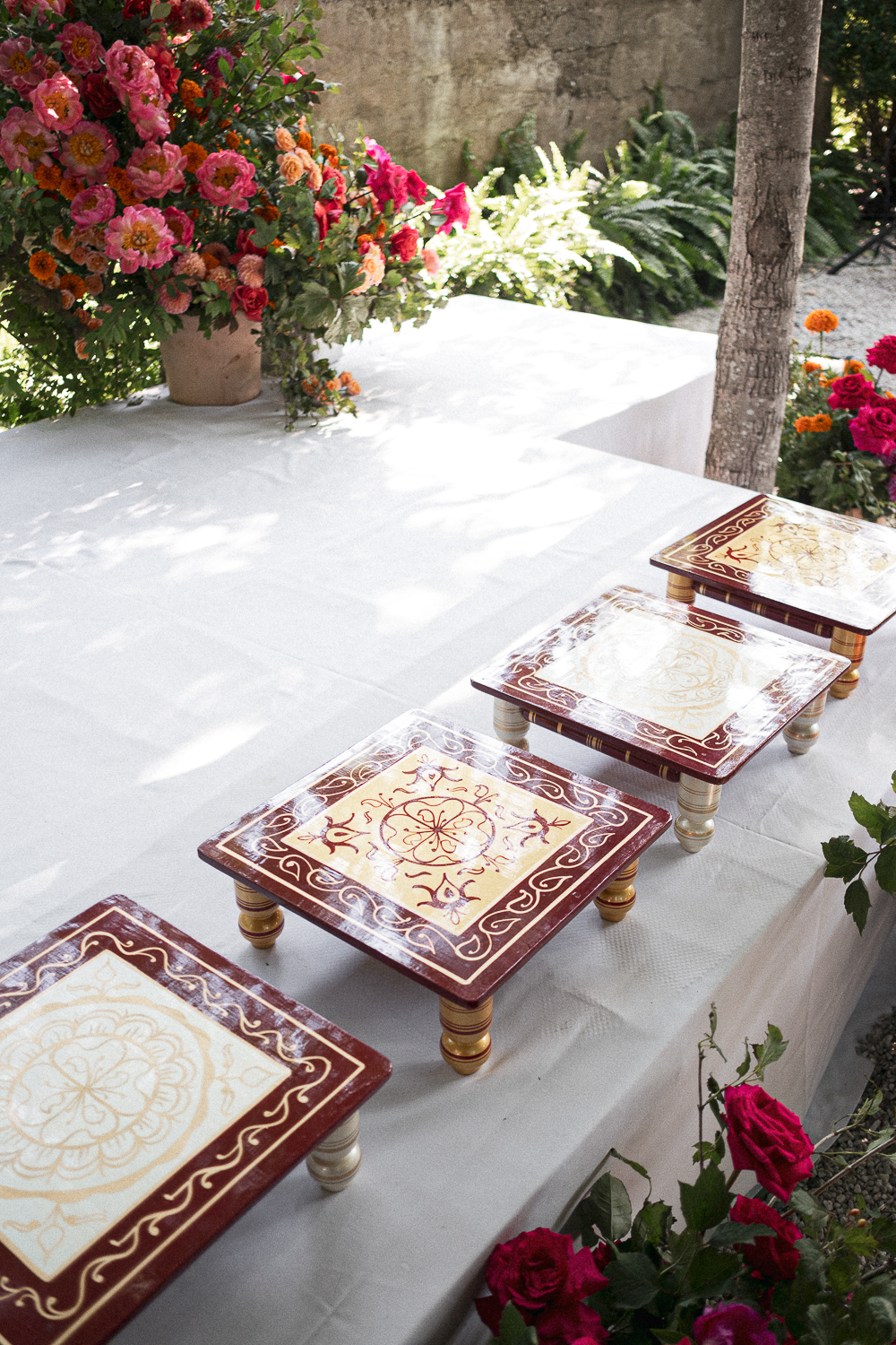 A white table with decorative tiles and a large floral arrangement in a pink pot, surrounded by greenery and flowers including roses and marigolds, outdoors in a garden setting.