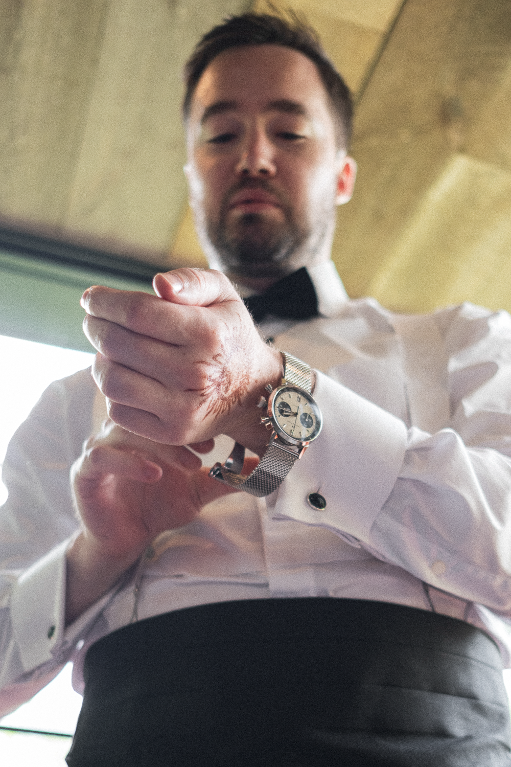 A man in formal attire, including a white shirt with cufflinks and a black bow tie, looking at his watch while adjusting it.