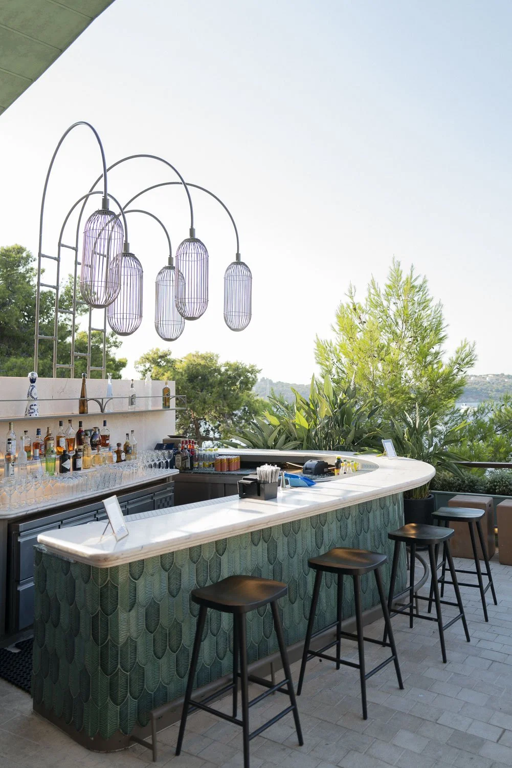 Outdoor bar with a white marble countertop, black stools, and a curved green mosaic base, set under decorative hanging lights, overlooking trees and a blue sky.