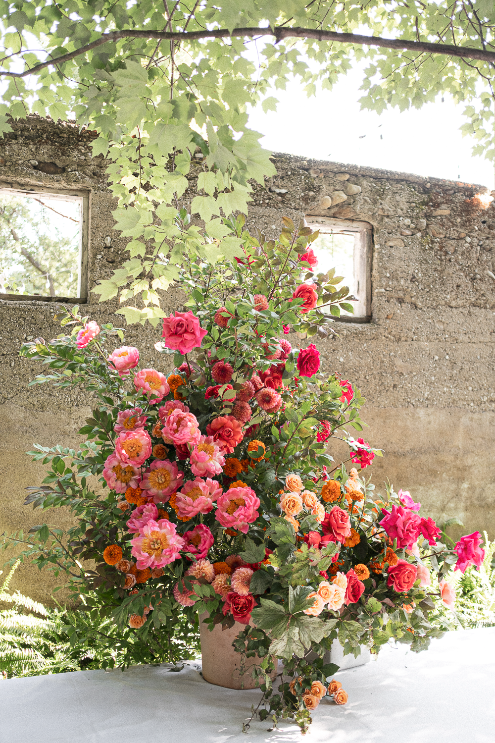 Colorful flower arrangement with pink, red, orange, and peach roses and other flowers in a large beige vase on a table in front of a rustic stone wall with small windows and green leafy plants.