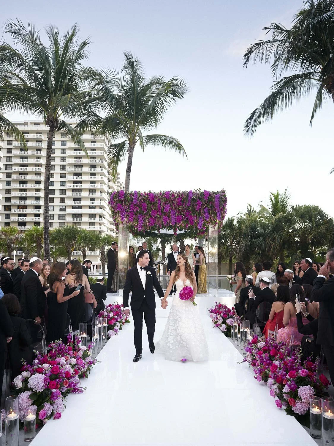 A newlywed couple walks down the aisle at an outdoor wedding ceremony, surrounded by guests, with palm trees, flowers, and a tall building in the background.
