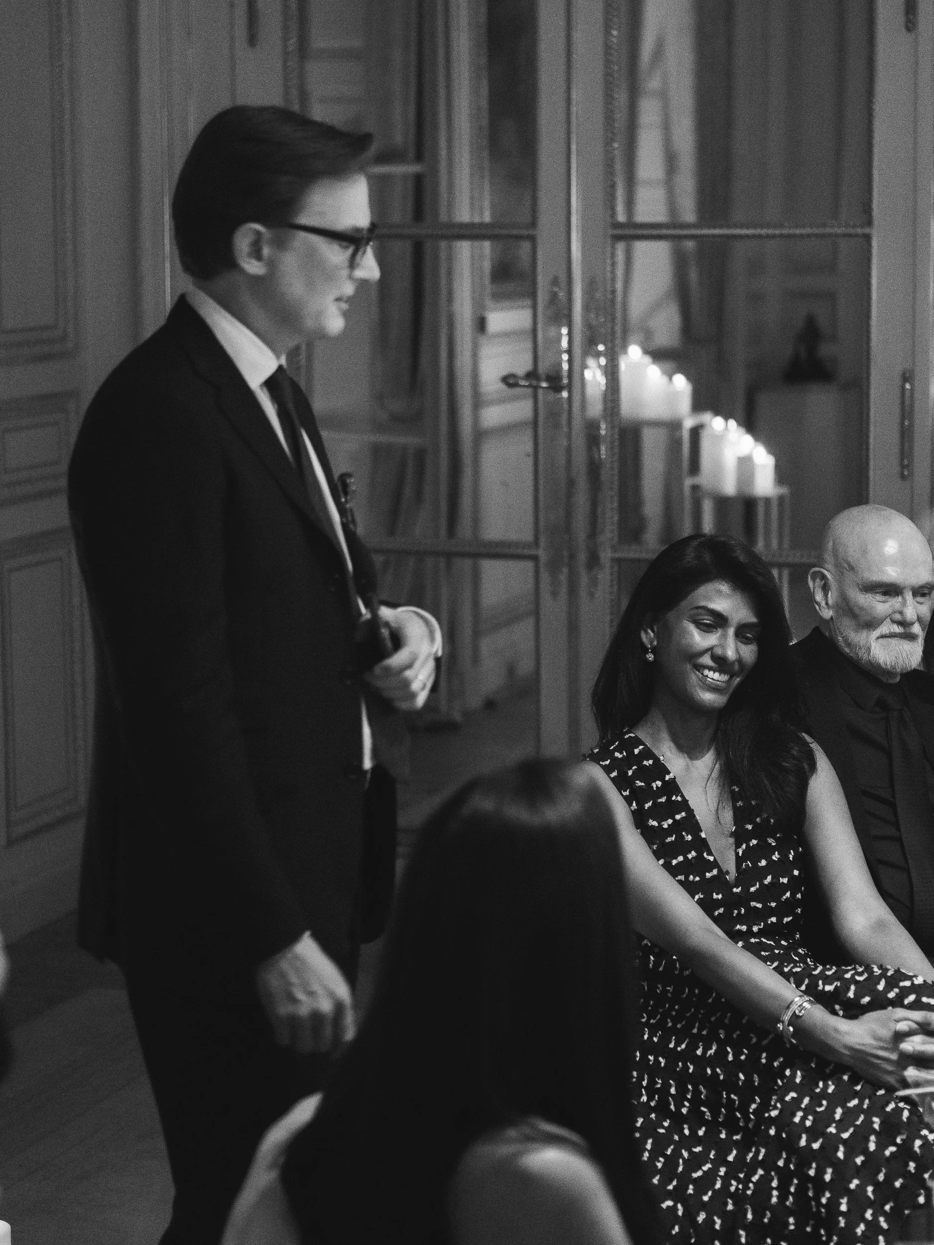 A black and white photo of four people sitting at a table during a formal gathering. One man stands, speaking or listening, while three others are seated. They are indoors with candles on the wall in the background.