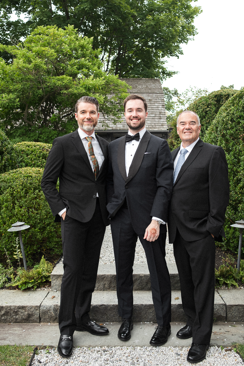 Three men in suits posing outdoors on stone steps, greenery, and trees in the background.