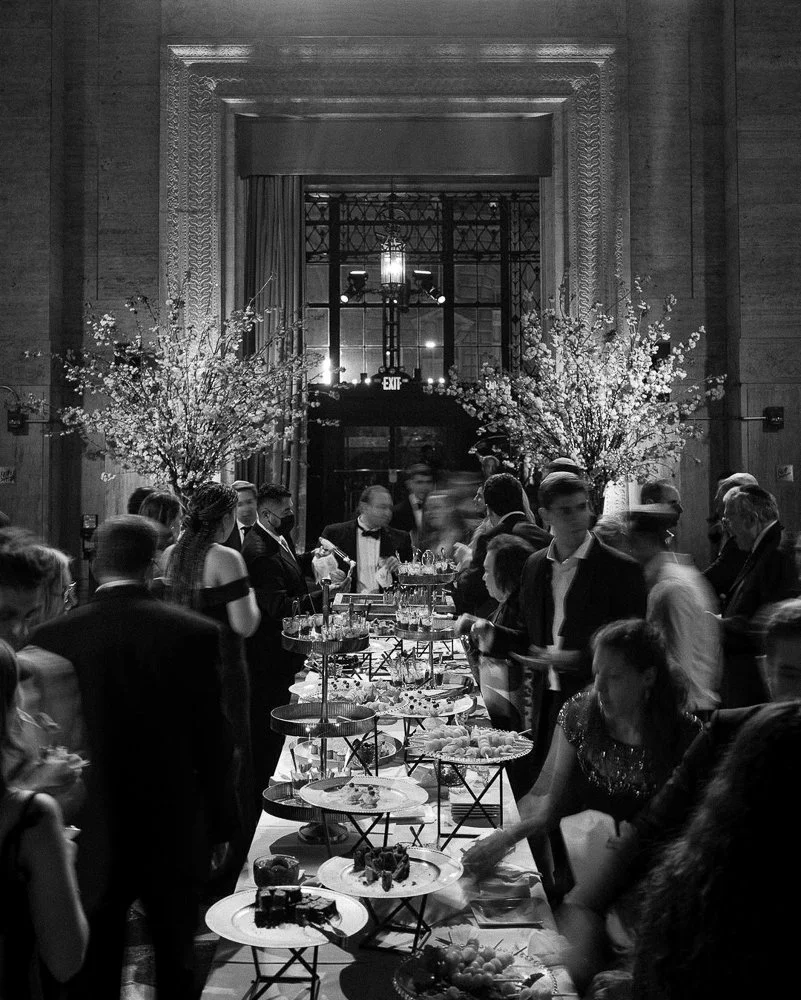 Black and white photo of a formal gathering at a long buffet table with desserts, surrounded by guests in tuxedos and formal dresses, in an elegant room with large windows and floral arrangements.