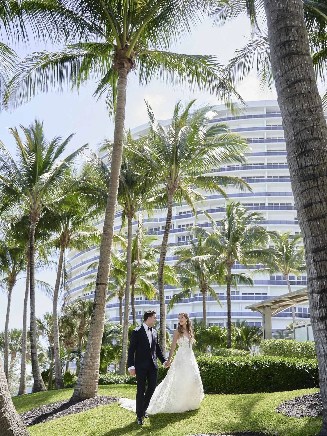A bride and groom walking hand in hand in a lush, tropical garden with tall palm trees and a modern, curved building in the background.
