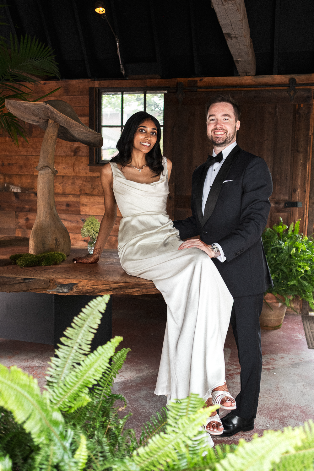 A couple dressed in formal wedding attire: the woman in a white gown and the man in a tuxedo, sitting on a wooden table in a rustic indoor setting decorated with plants and a large mushroom sculpture.