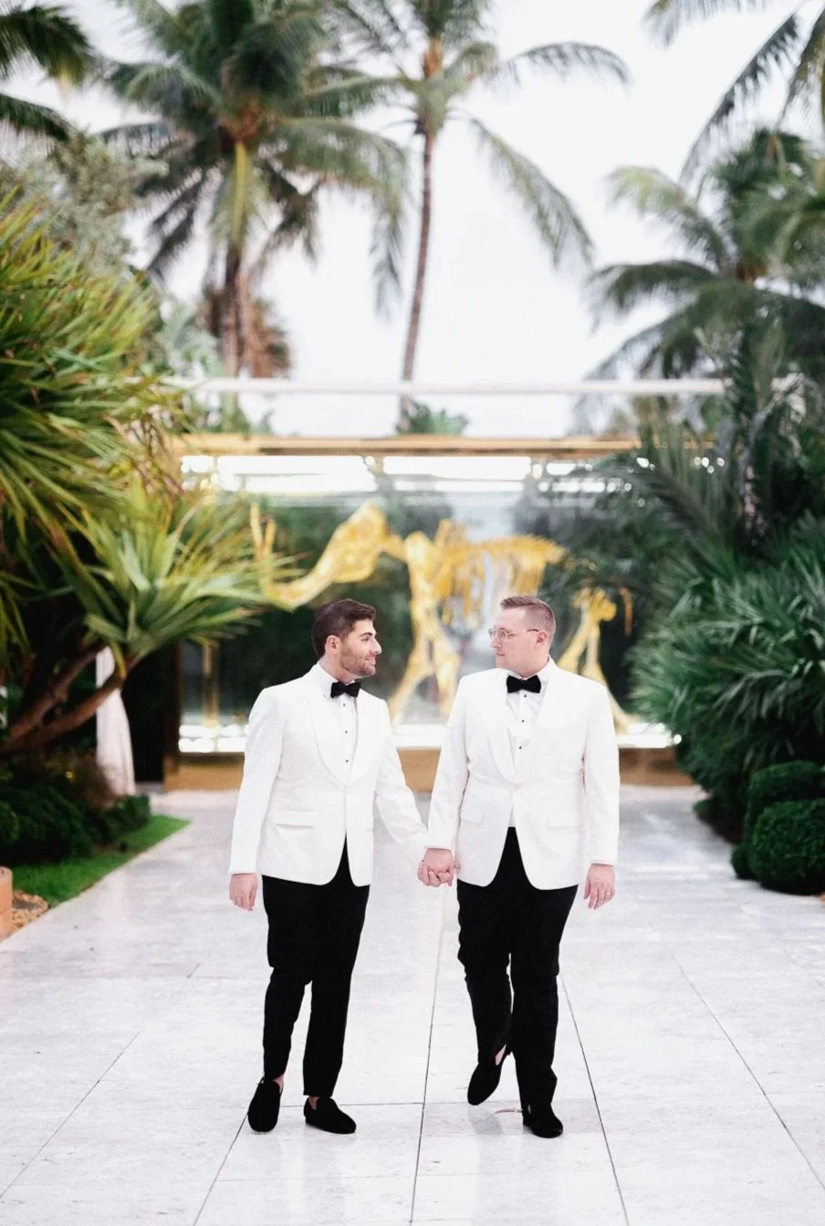 Two men dressed in white tuxedo jackets, black pants, and bow ties holding hands and walking outdoors in front of lush greenery and tall palm trees.