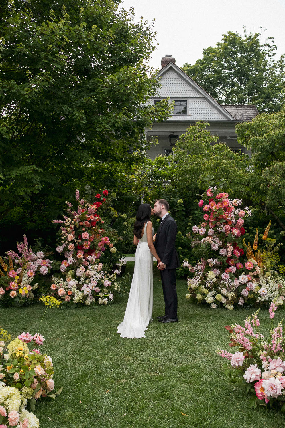 A bride and groom kiss in a garden surrounded by colorful flowers, with a house and trees in the background.