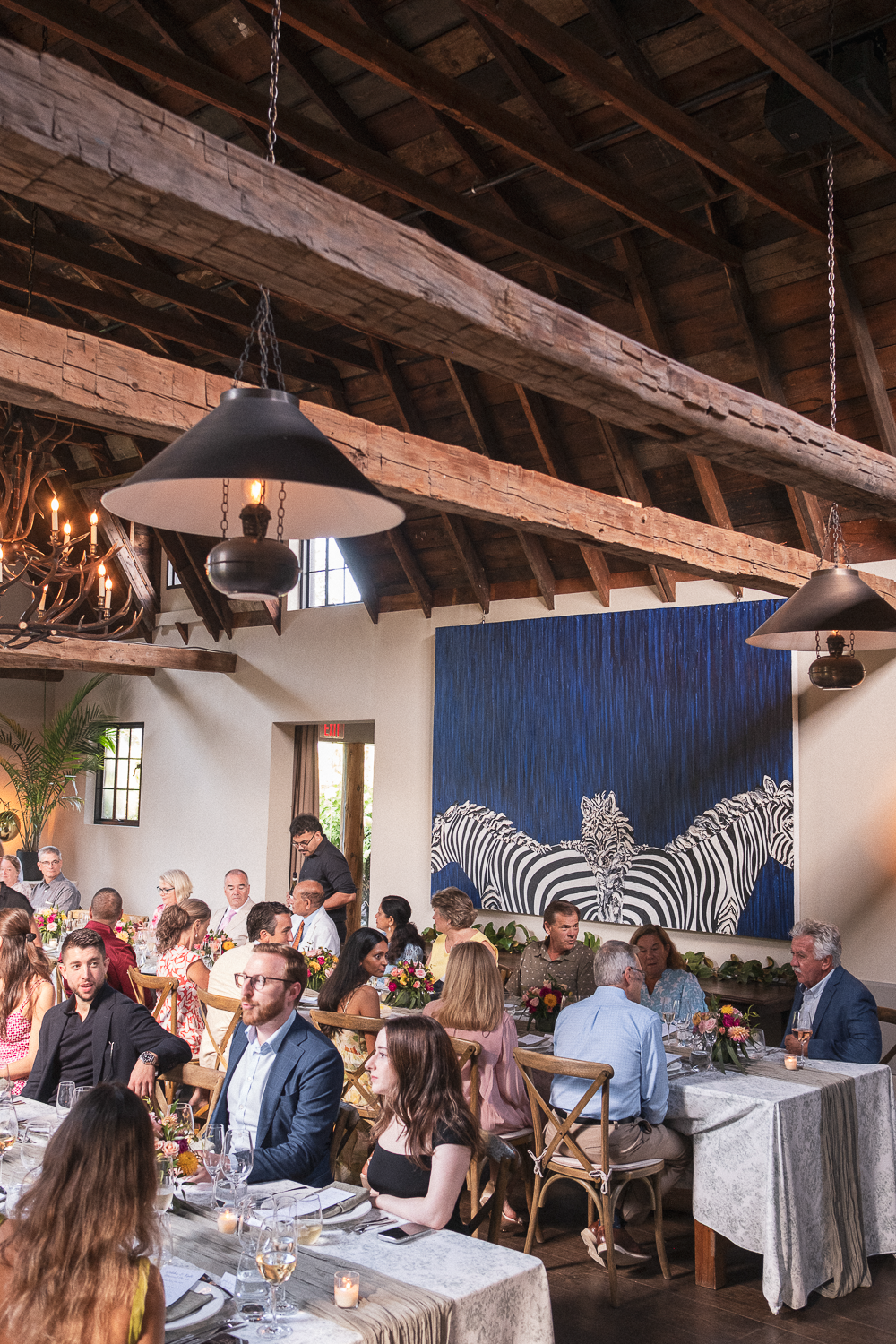 People seated at tables during a formal event or dinner in a rustic restaurant with wooden beams and a large zebra artwork on the wall.