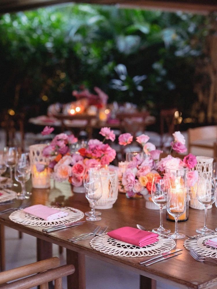 A tablescape with pink and peach floral arrangements, pink napkins, glassware, and candle lighting for a celebration or event.