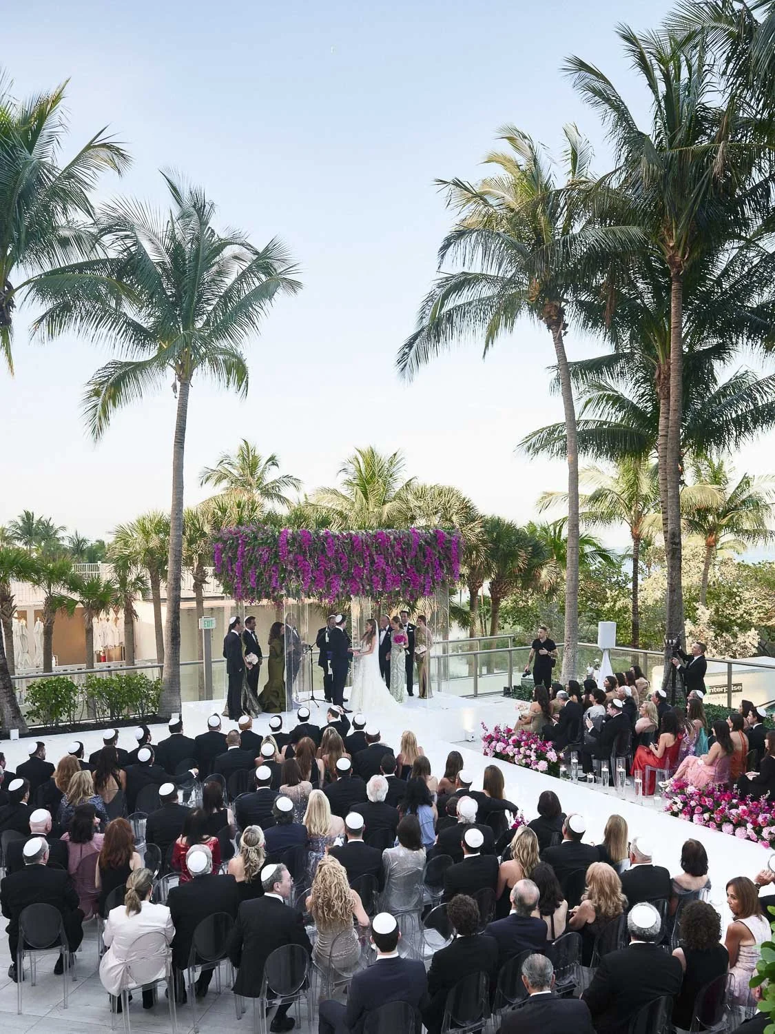 Wedding ceremony outdoors under palm trees with a large crowd of guests.