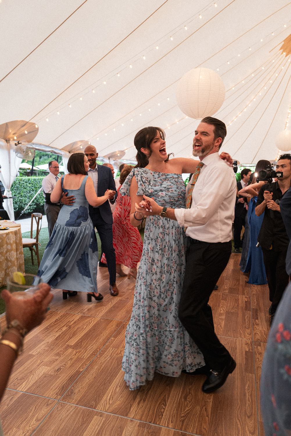 People dancing and celebrating under a decorated tent at a wedding reception, with string lights, paper lanterns, and guests in colorful dresses and suits.