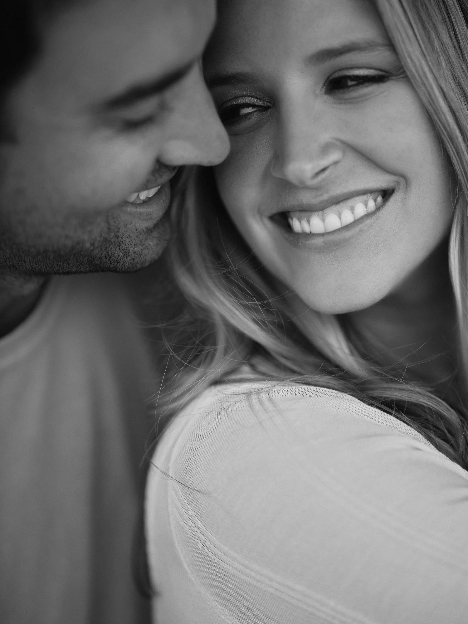 Close-up black and white photograph of a smiling woman with straight hair and a man with facial hair. The image depicts their faces closely together, with the woman's face turned slightly towards the camera and the man's face partially visible.