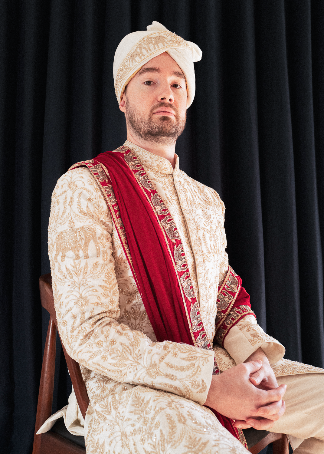 A man in traditional South Asian wedding attire, wearing an elaborate cream-colored sherwani with gold embroidery, a matching turban, and a red sash draped over his shoulder, seated against a black curtain background.