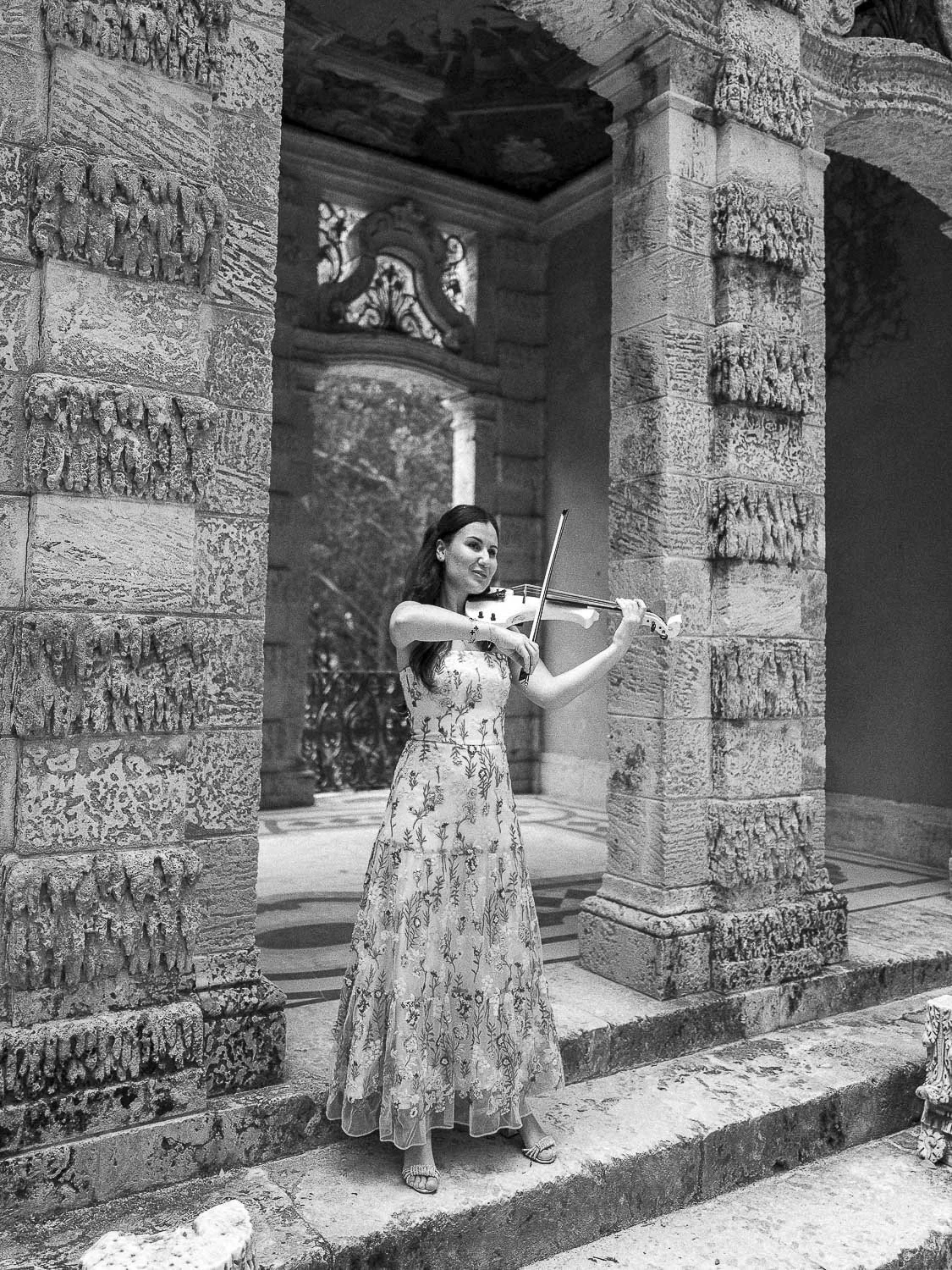 A woman in a floral dress playing a violin on stone steps in front of a historic stone building with ornate architectural details.