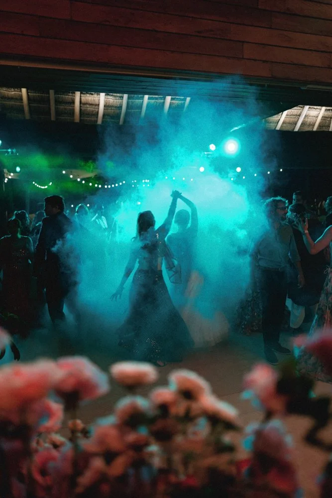 People dancing at a party or celebration with colorful lights and fog, under a wooden ceiling.