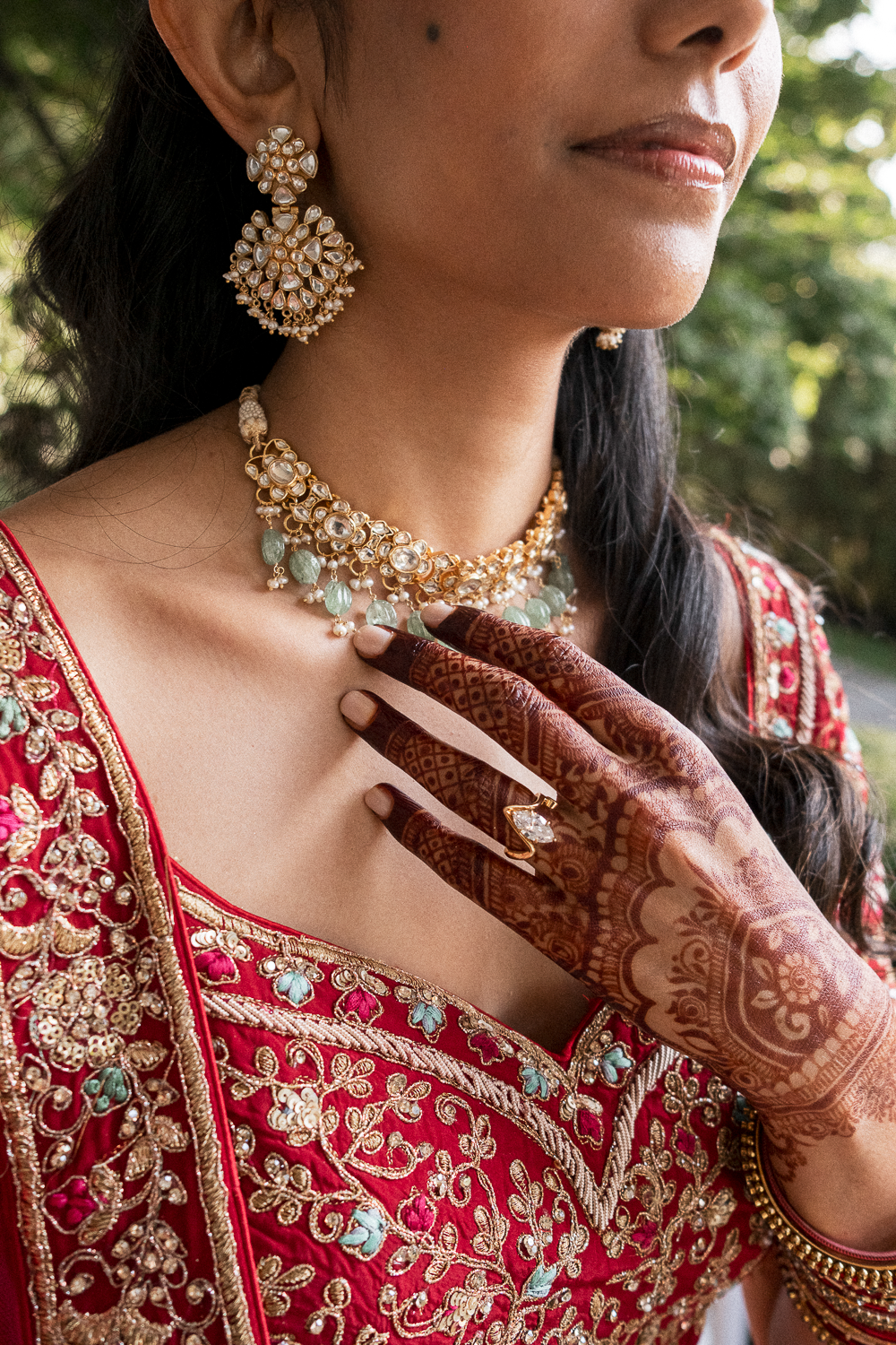 Close-up of a woman wearing red and gold embroidered traditional attire, adorned with gold and pearl jewelry, seen outdoors with blurred green foliage in the background.