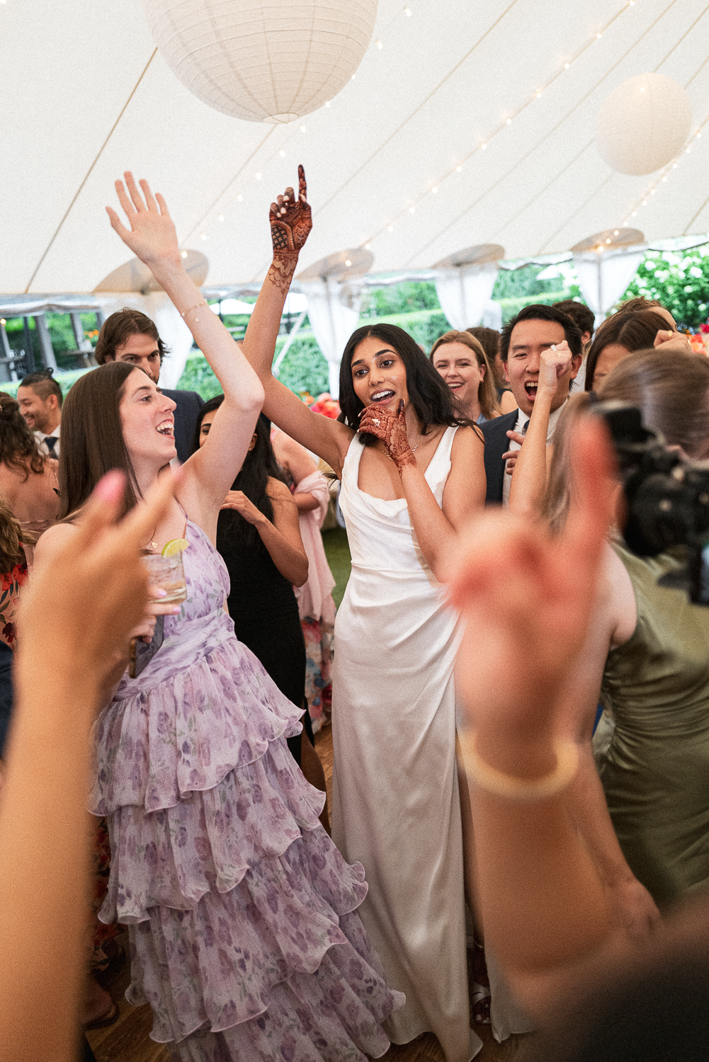 People dancing and celebrating at a wedding reception inside a tent, with some wearing formal attire, including a woman in a white dress and another in a purple tiered dress.