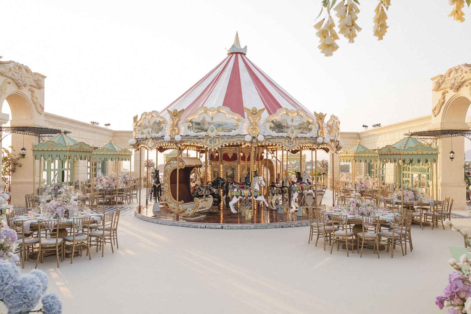 Decorative carousel with painted horses surrounded by pastel-colored tables and chairs, floral centerpieces, and ornate buildings on a sunny day.