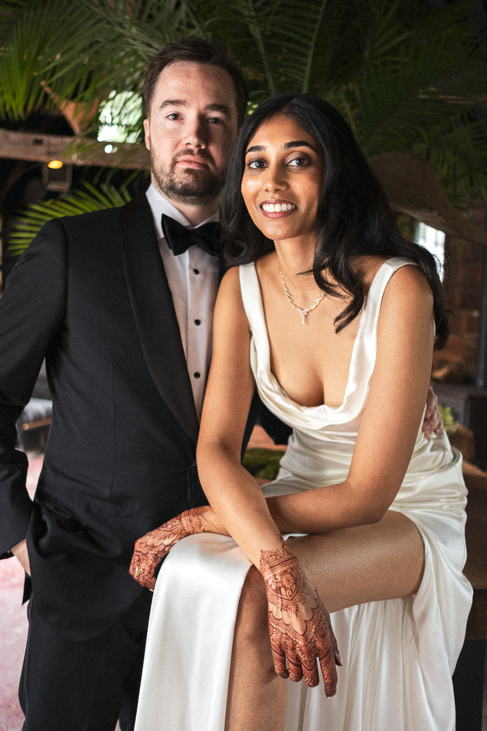 A man in a tuxedo and a woman in a satin dress, both dressed for a formal event, posing indoors with tropical plants in the background.