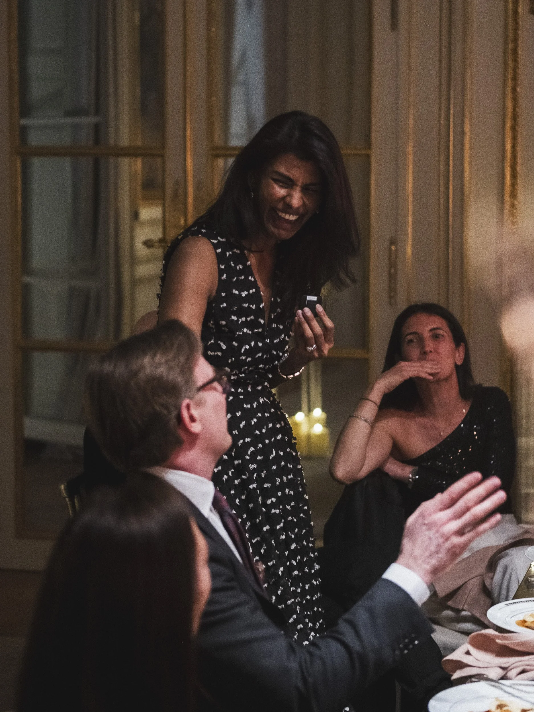 A woman standing and laughing with a group of people at a dinner party in an elegant room with gold framed mirrors.