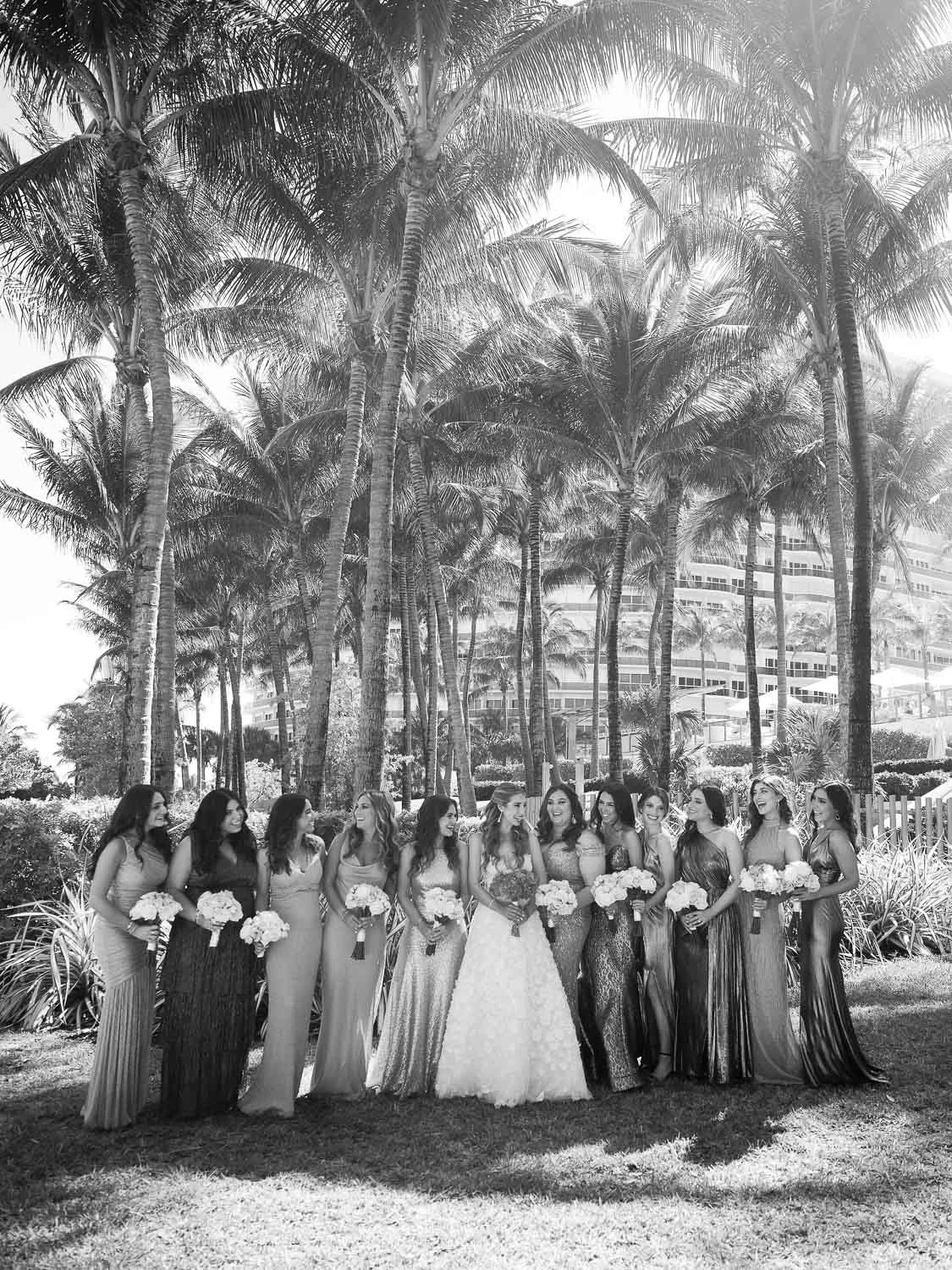 A group of women in formal dresses standing outdoors among tall palm trees, holding bouquets, with a high-rise building in the background.