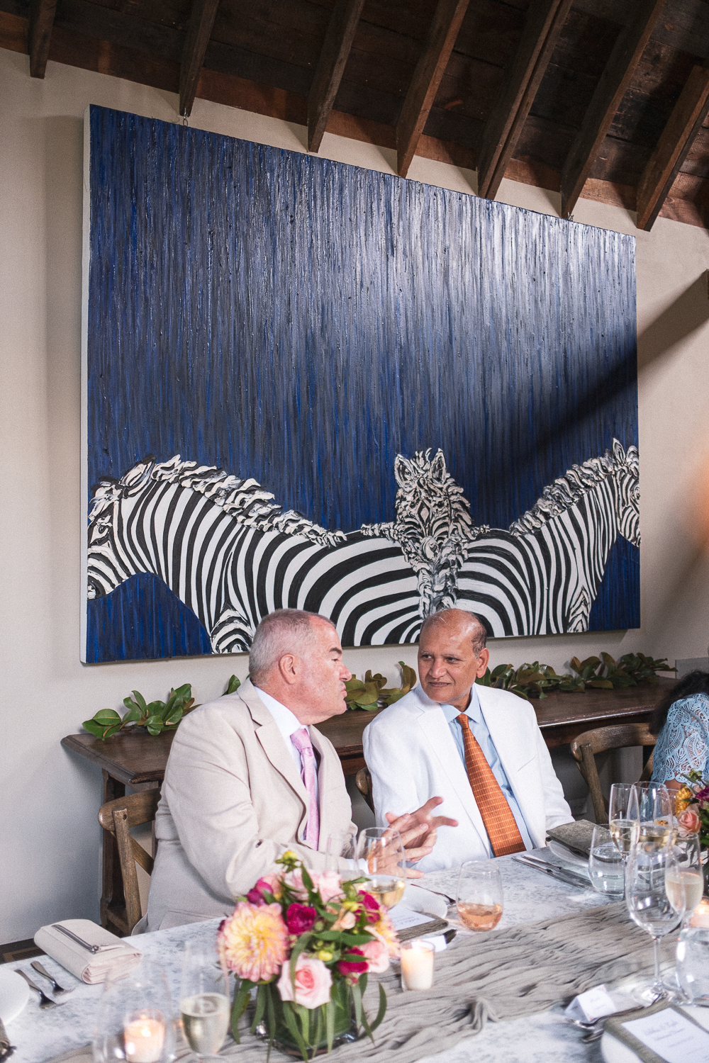 Two men in suits sitting at a table engaged in conversation during a formal gathering or dinner, with a large wall art depicting zebras beneath a blue sky in the background.