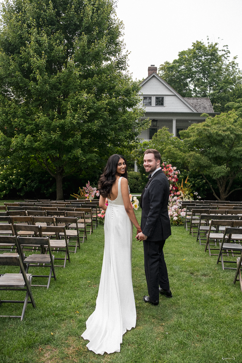 A bride and groom holding hands and smiling during their outdoor wedding ceremony in a garden, with rows of empty chairs, lush green trees, and a white house in the background.