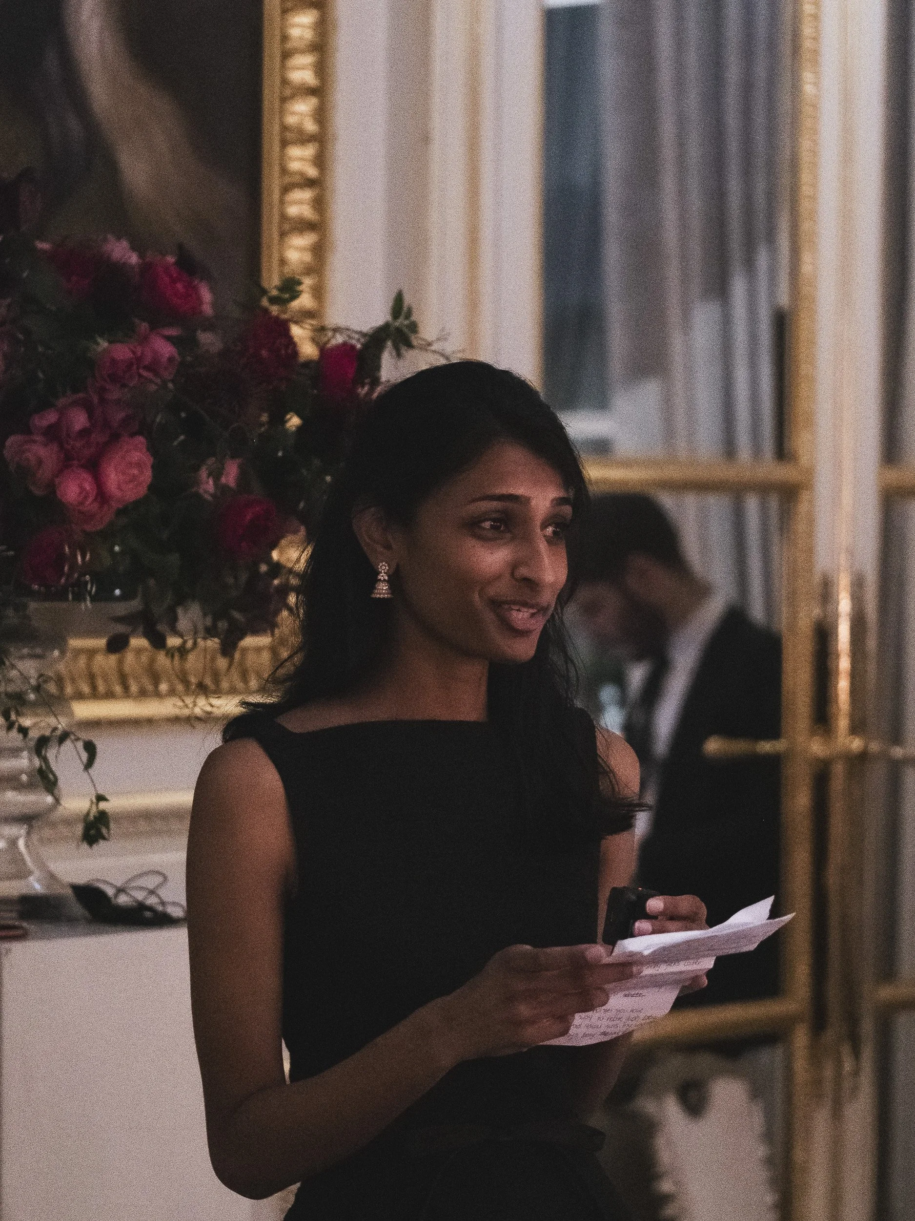 A woman in a black dress standing indoors, holding notes or a speech, with an ornate gold mirror and pink floral arrangement in the background.