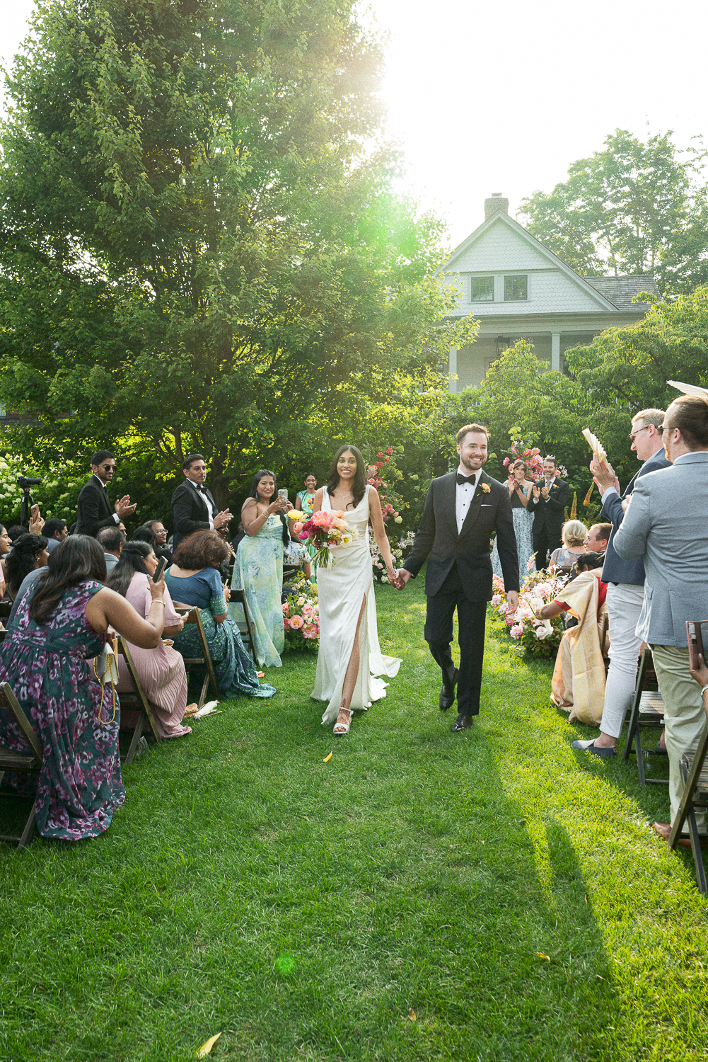 A newlywed couple walking hand in hand down the aisle at their outdoor wedding ceremony, surrounded by friends and family, with a house and trees in the background.