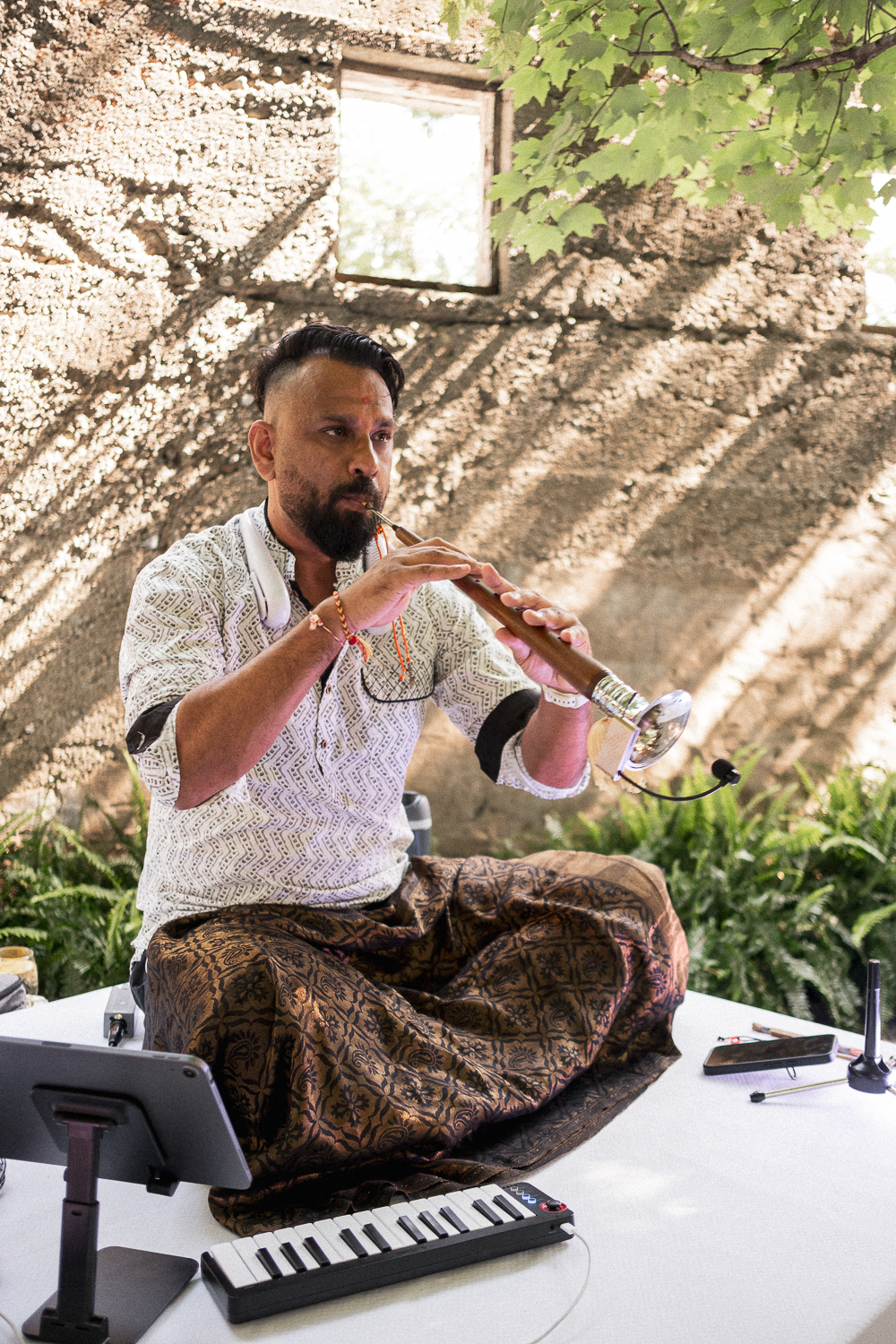 A man sitting cross-legged on a white table, playing a flute in a natural indoor setting with sunlight streaming through an open window and green leaves overhead.