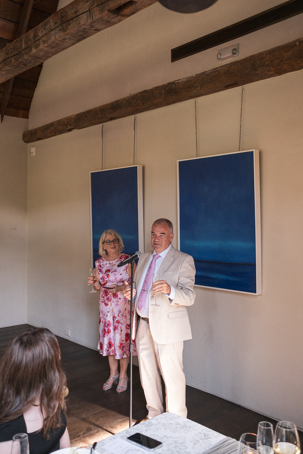 A man in a beige suit and pink tie speaking at a microphone during an event, with a woman in a pink floral dress holding a glass of wine standing behind him. There are two large blue artwork pieces hanging on the wall behind them, and an audience mem