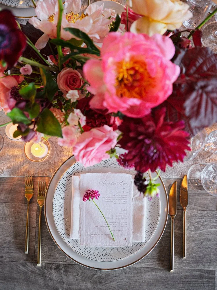 A table setting with a floral centerpiece, a dinner plate, gold utensils, a linen napkin with a flower illustration and text, a candle, and wine glasses on a wooden table.