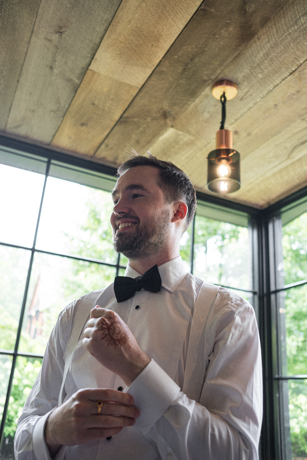 A smiling man with a beard wearing a white dress shirt and black bow tie, adjusting his shirt cuff, indoors with large windows and a wooden ceiling, natural light, greenery outside.