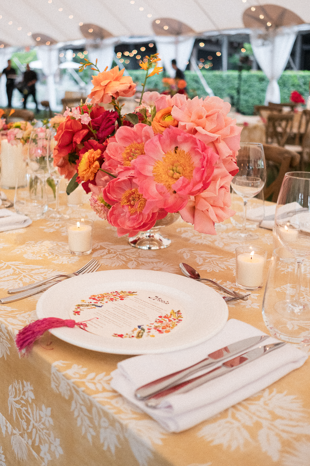 A table setting with a large pink floral centerpiece at an event, with candles, glassware, and a printed menu on a white plate, under a white canopy with outdoor seating visible in the background.