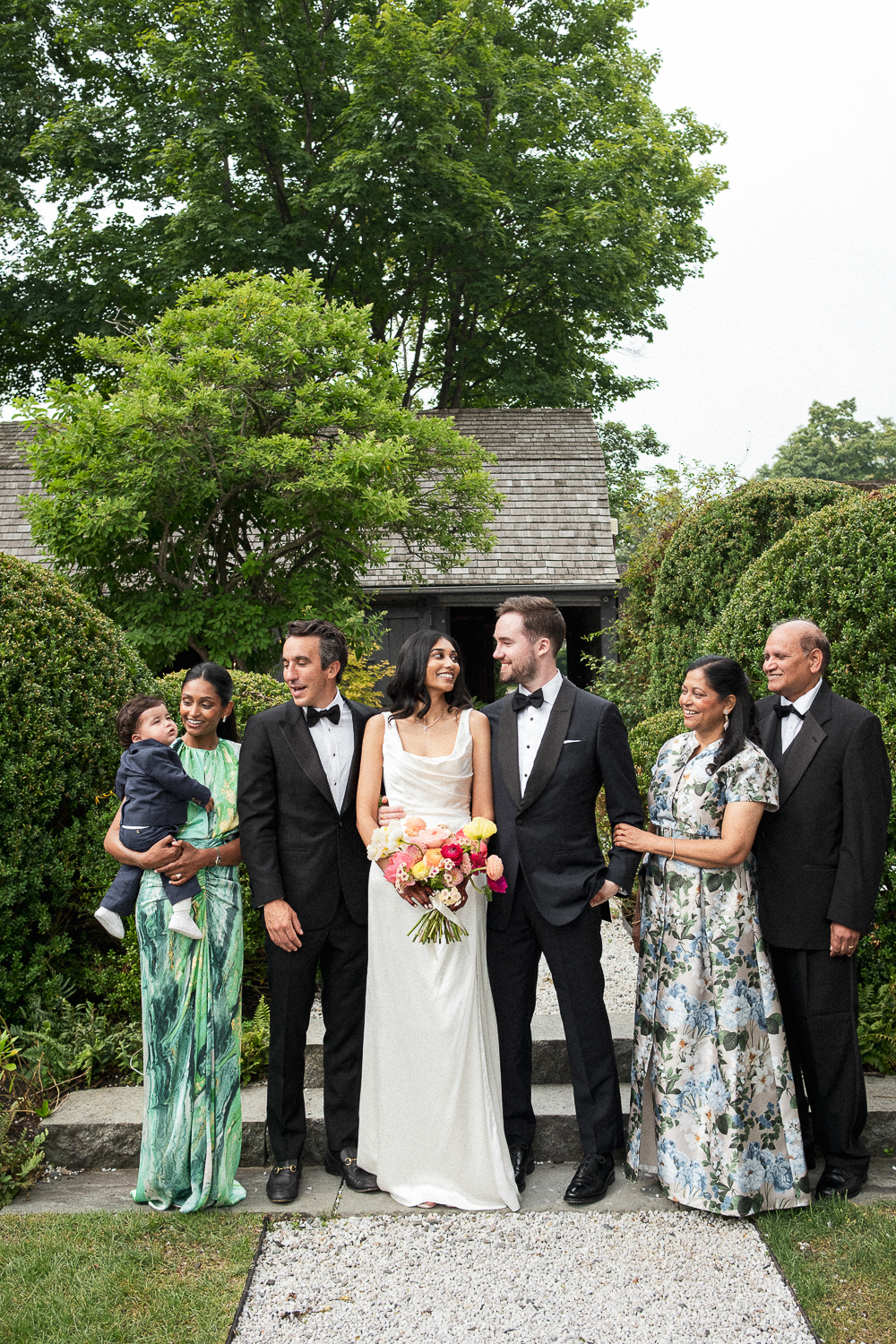 A wedding party of seven people standing on a garden path, with a small building and leafy trees in the background. The bride holds a bouquet, wearing a white dress, with dark hair. The groom and another man are in tuxedos. Four other adults, includi
