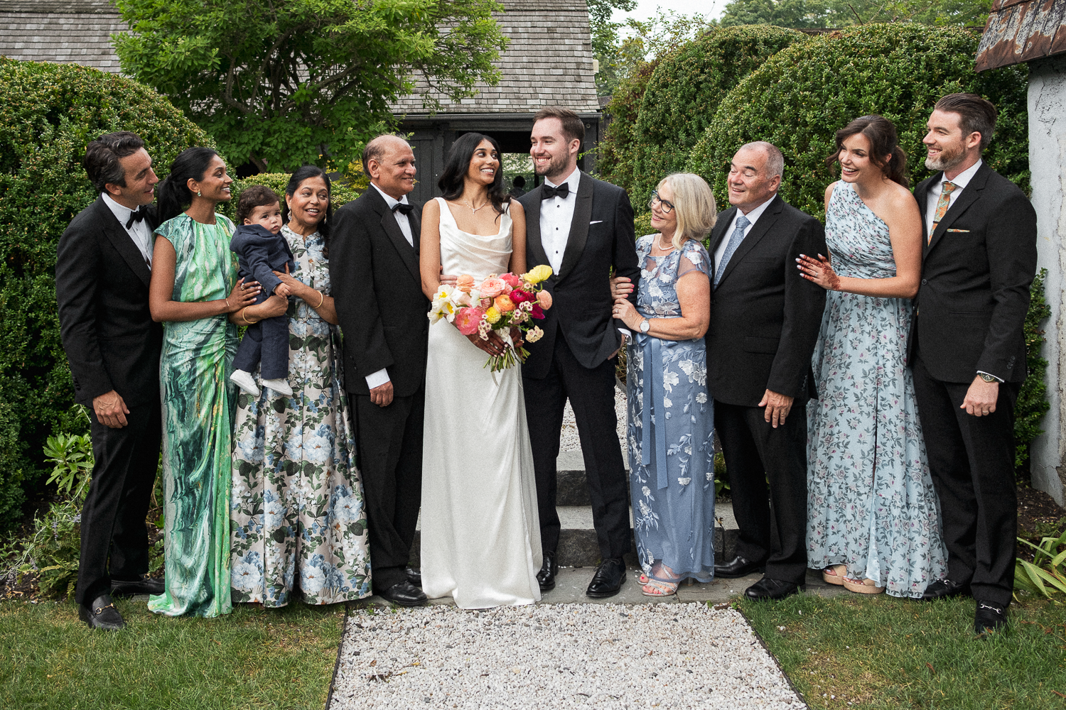 A wedding party outdoors with the bride and groom in the center, surrounded by family and friends. The bride is holding a bouquet of colorful flowers, and everyone is dressed in formal attire, smiling and celebrating.
