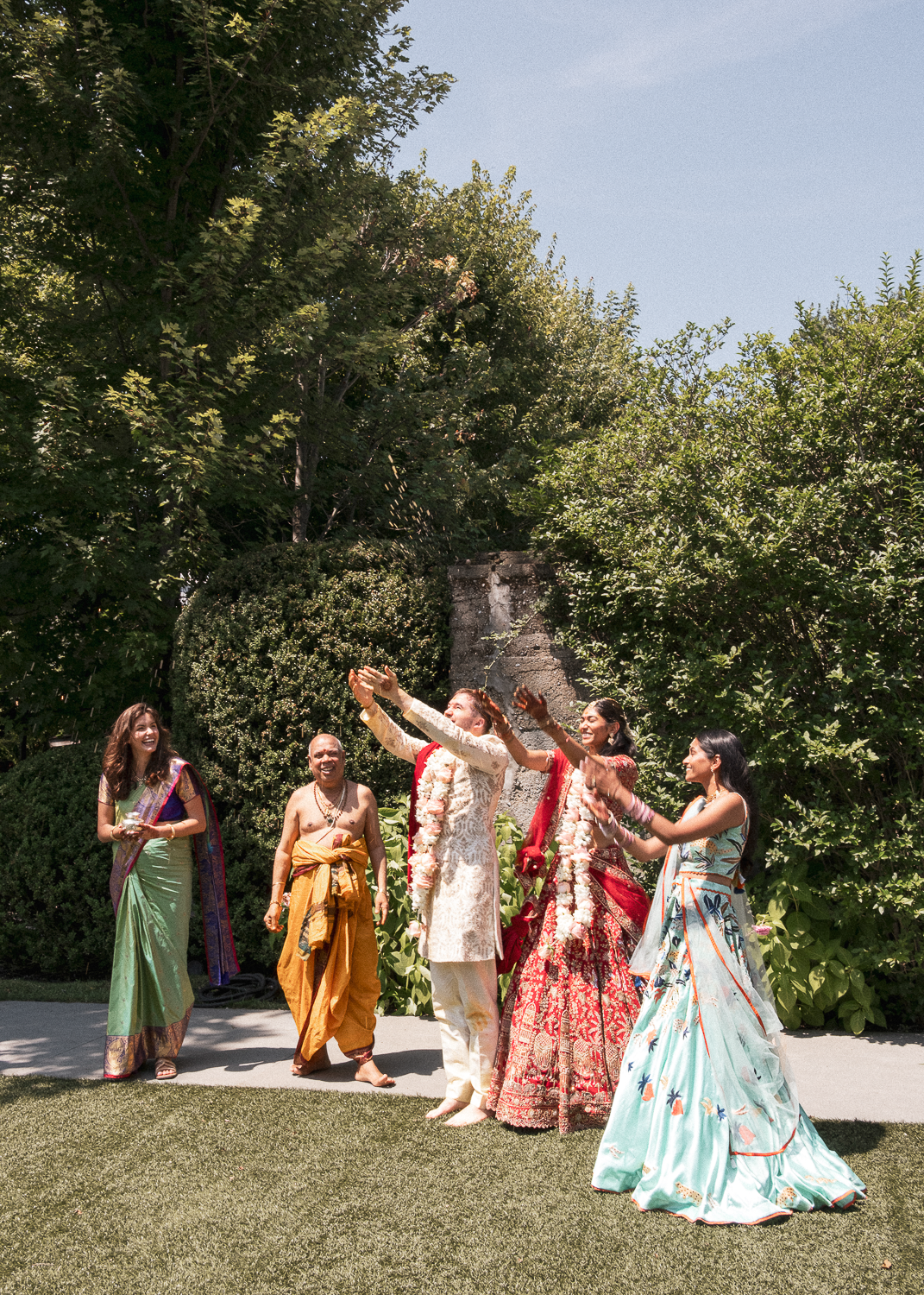 A group of five people dressed in traditional Indian wedding attire celebrating outdoors under a clear sky with trees and bushes behind them.