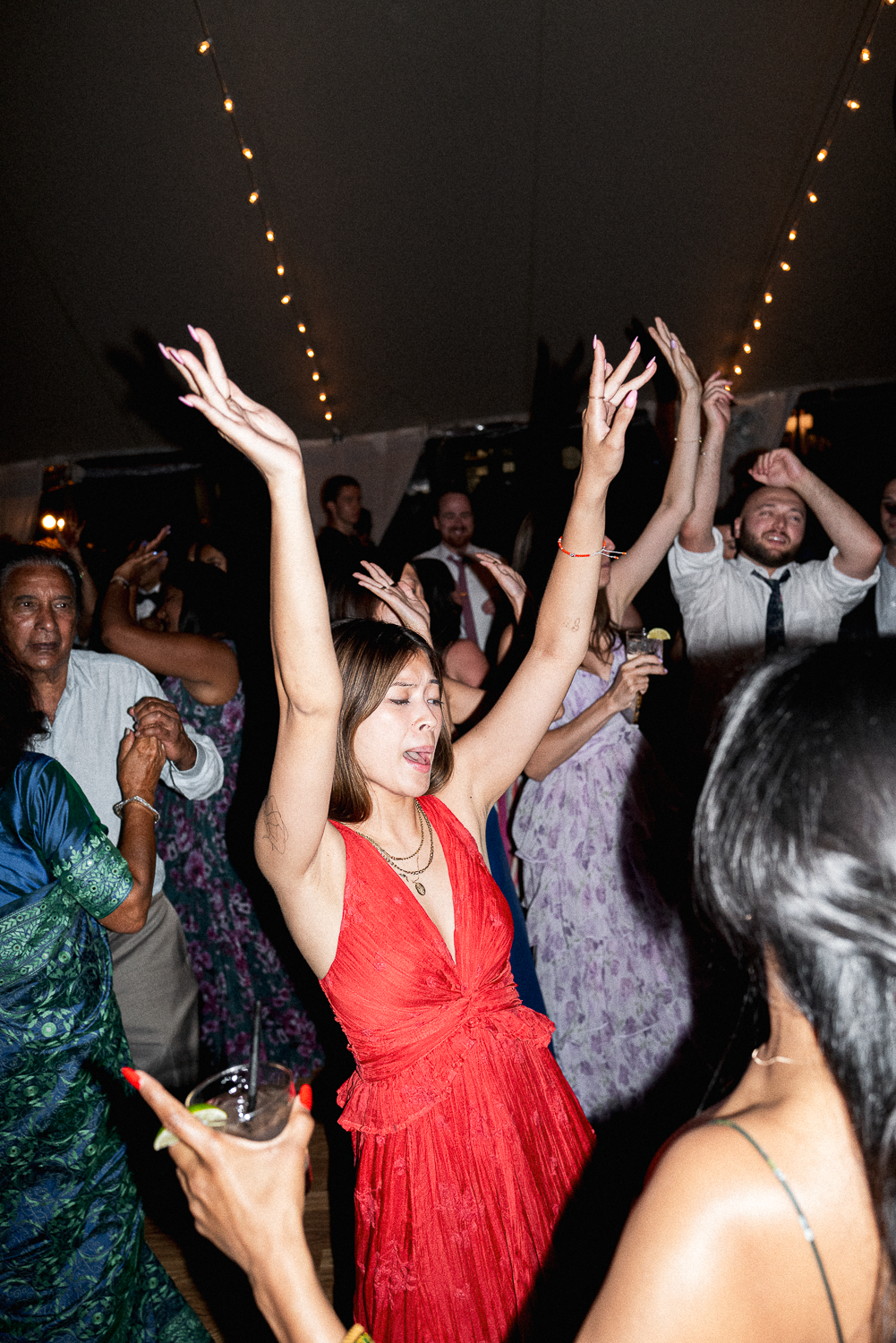 People dancing and celebrating at a party or wedding under string lights, with a woman in a red dress in the foreground raising her hands and with a joyful expression.