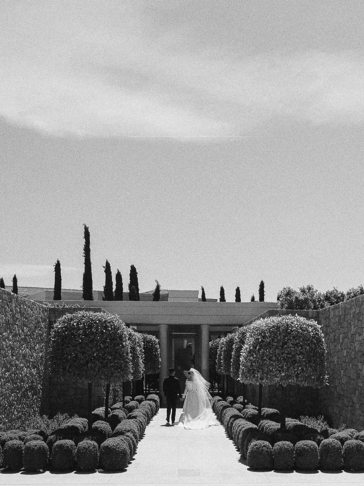 Black and white photo of a wedding couple walking down a garden pathway lined with trimmed bushes and trees, with tall cypress trees in the background under a sky with scattered clouds.