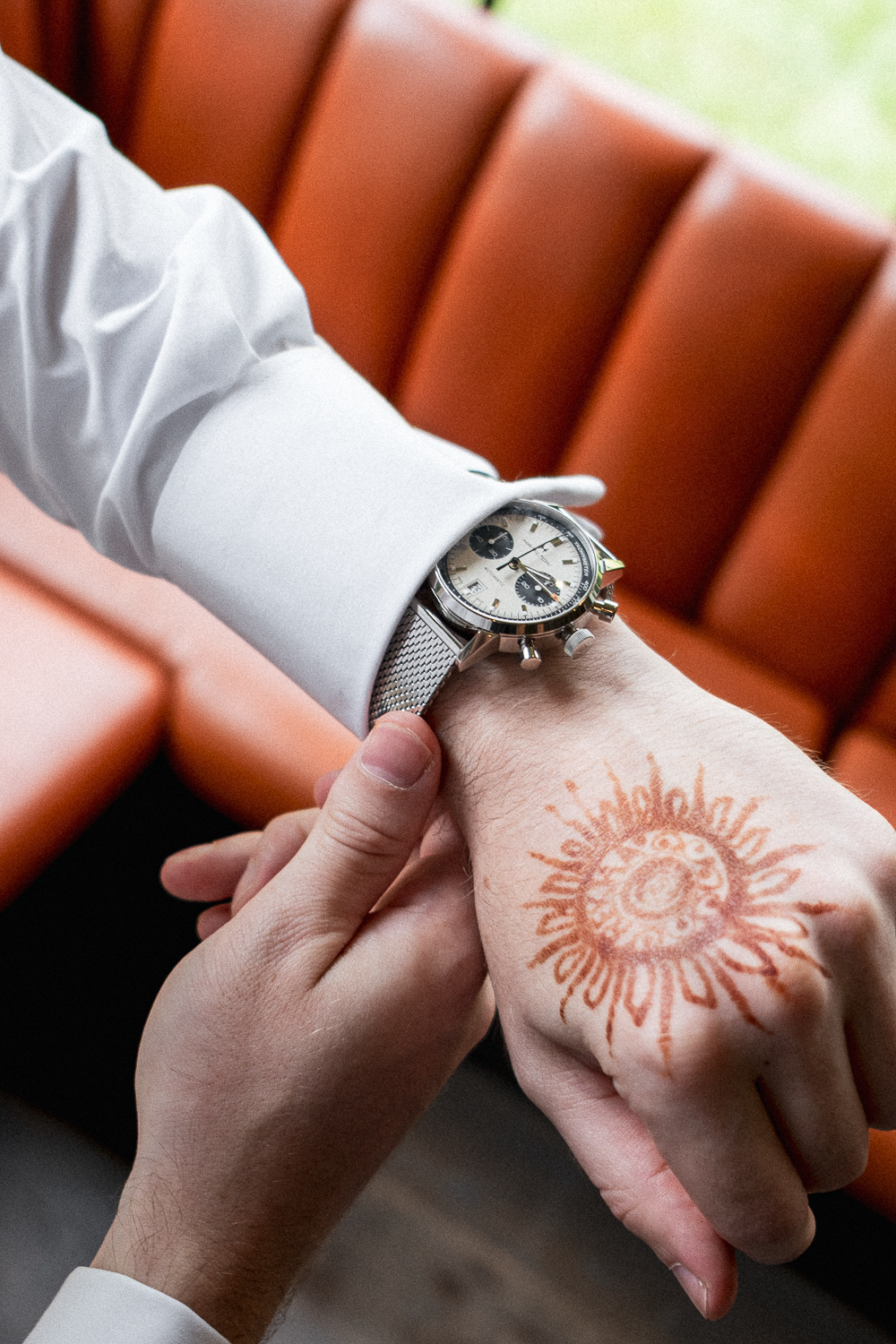 A person is checking the time on their wristwatch while another person touches their wrist. The person wearing the watch has a sun-shaped tattoo on their hand. The setting includes orange benches or chairs and a window with natural light.