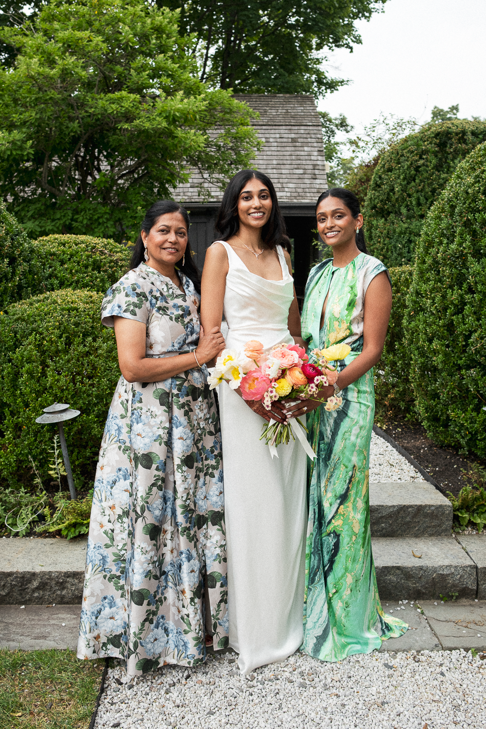Three women standing outdoors among greenery, with one woman in the center holding a bouquet of flowers, wearing a white dress, flanked by women in floral and green dresses.